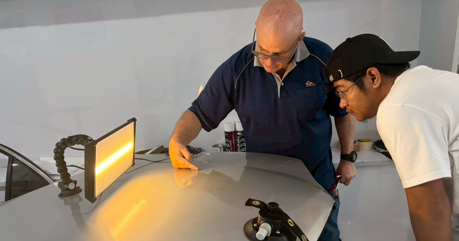 A trainer points at a grey training panel while a learner watches. A yellow PDR lamp stands above the panel, casting a broad reflection on its surface.