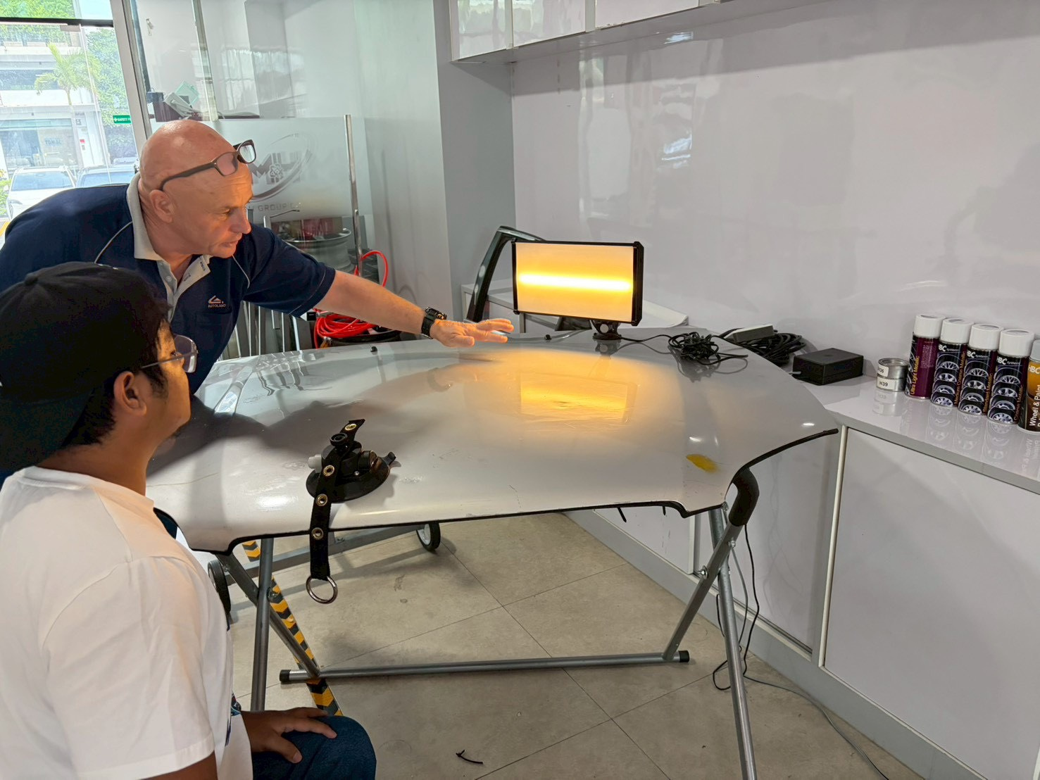 A seated learner and a trainer view a panel secured on a stand. The trainer gestures above a warm reflection cast by the lamp at the far edge of the panel.